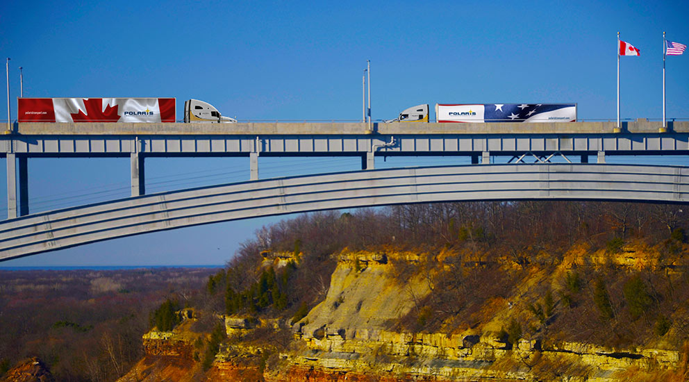 One Polaris truck with Canadian trailer driving toward another Polaris truck with U.S. trailer on bridge 