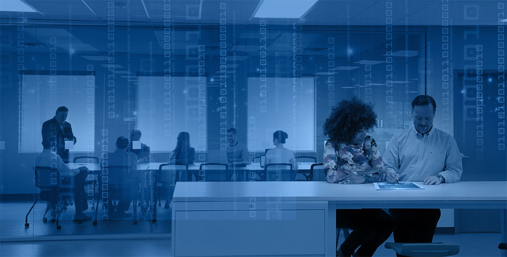 Office with two people sitting at desk and group in conference room in background 