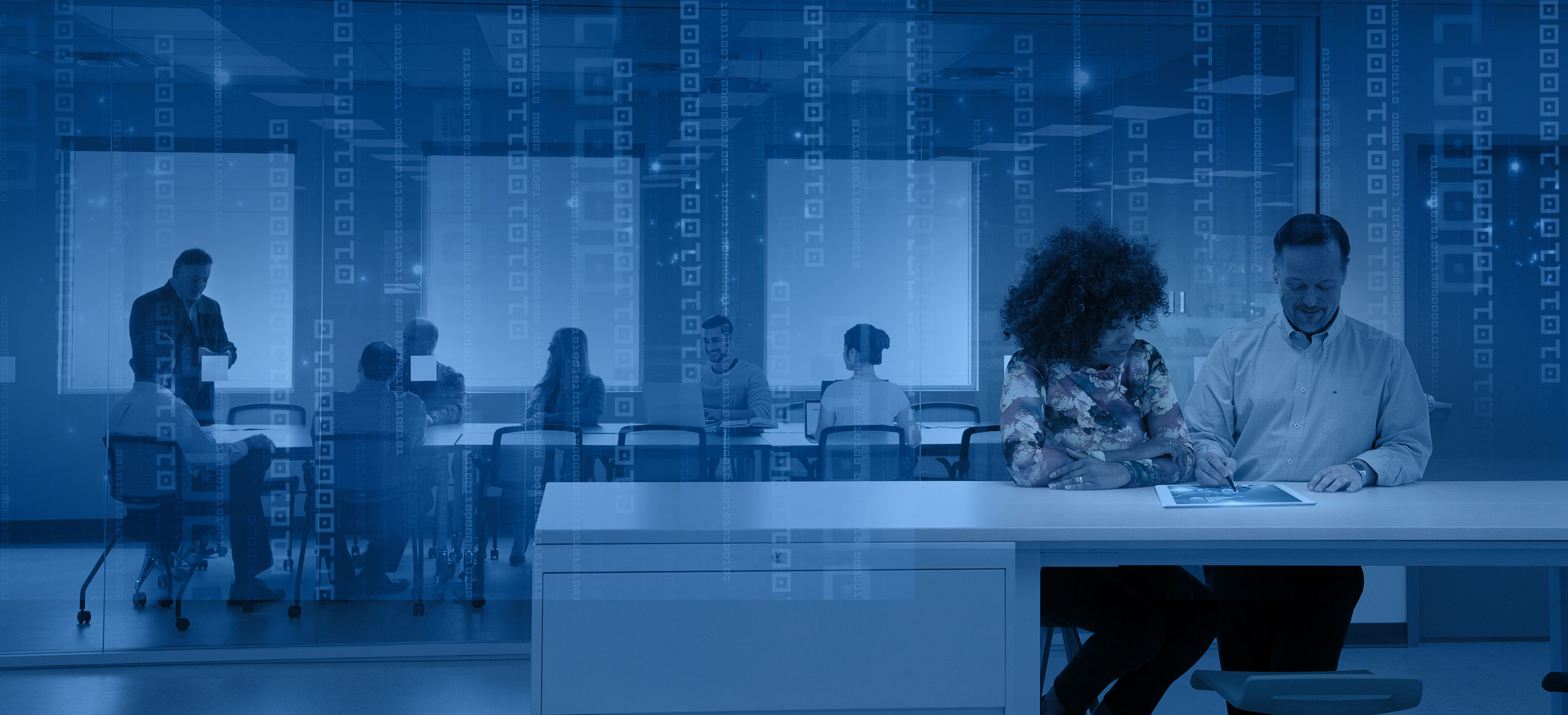 Office with two people sitting at desk and group in conference room in background 